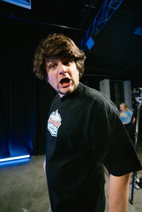 A man with curly brown hair wearing a black graphic t-shirt makes an intense, wide-eyed facial expression in what appears to be a backstage studio setting