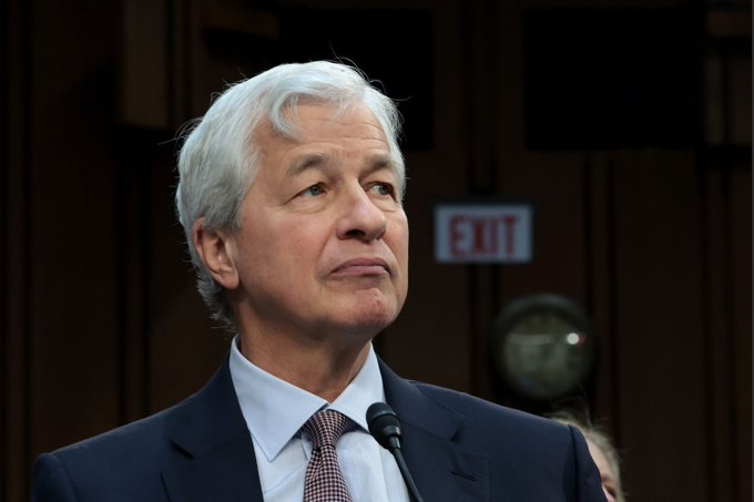 A close-up portrait of a man with gray hair wearing a dark suit and patterned tie, speaking at a microphone in a formal indoor setting with an exit sign visible in the background.