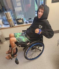 A young person sitting in a wheelchair on an indoor patio, wearing a black hoodie and green shorts, giving a thumbs-up gesture.