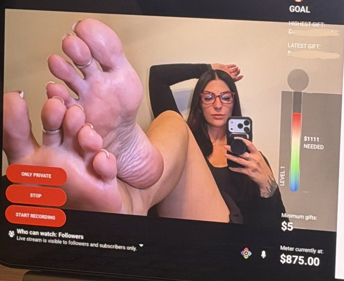 A photograph of a screen showing an adult webcam stream featuring a woman holding her feet up to the camera while taking a mirror selfie, with on-screen controls including "ONLY PRIVATE," "STOP," "START RECORDING," and a meter showing "$875.00" toward a goal of "$1111 NEEDED."