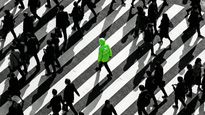 An aerial black-and-white photo of a crosswalk with one person in a bright green hoodie standing out among the monochrome crowd.