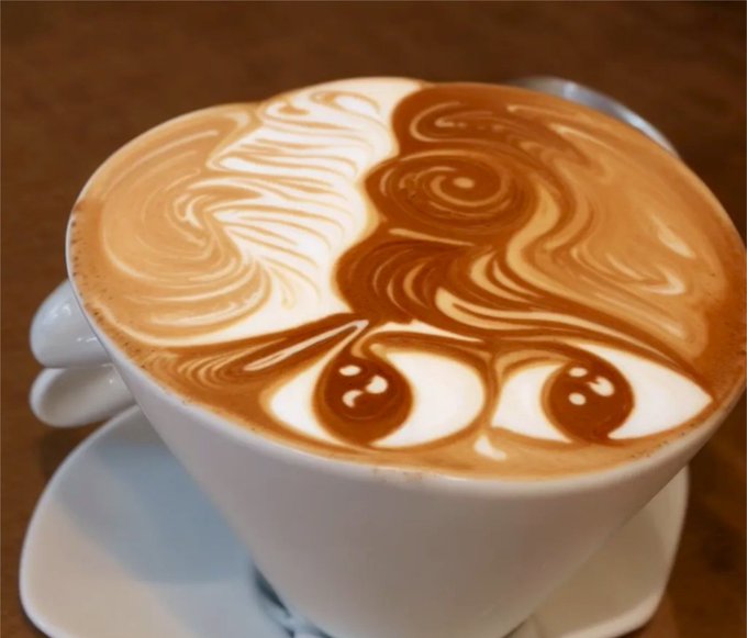 A latte featuring intricate latte art depicting a figure with flowing hair and cherries, served in a white cup on a saucer.