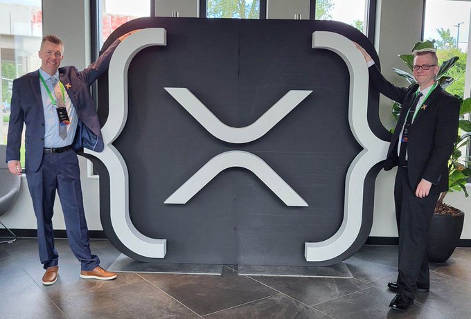 Two men in business suits stand on either side of a large, freestanding physical display of the XRP cryptocurrency logo inside a modern building with large windows.