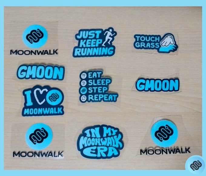 A collection of vinyl stickers scattered on a wooden surface featuring blue and black designs with text including 'MOONWALK', 'GMOON', 'JUST KEEP RUNNING', 'TOUCH GRASS', 'I <heart> MOONWALK', and 'IN MY MOONWALK ERA'.