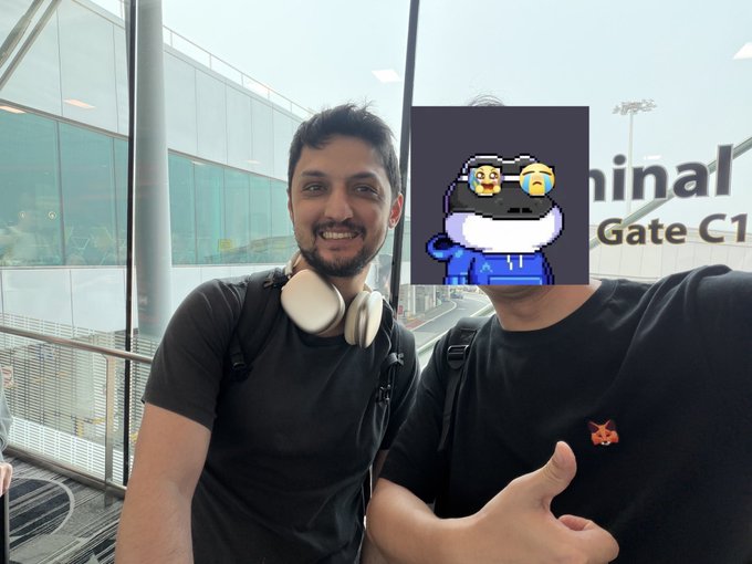 A selfie of two men at an airport terminal with text 'Gate C1' visible on a window. The face of the man on the right has been obscured with a cartoon penguin sticker featuring a crying emoji.