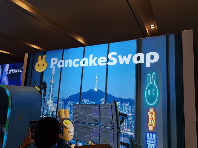 A PancakeSwap booth display at what appears to be a crypto conference, featuring the bunny logo, a cityscape backdrop, and trading screens.