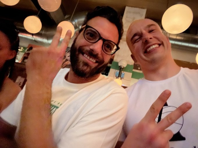 A selfie of two smiling men posing with peace signs—one with dark hair, glasses, and a beard, the other bald—both wearing white t-shirts in a warmly lit restaurant.