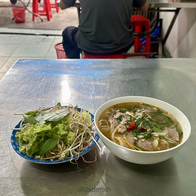 A photo of Vietnamese food on a stainless steel table featuring a bowl of beef noodle soup (pho) with herbs and chili, accompanied by a plate of fresh greens and bean sprouts, watermarked @0xdateth.
