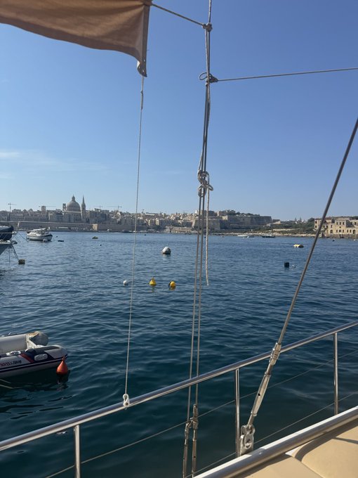 A scenic view from a sailboat in a Mediterranean harbor with a coastal city and domed church visible across the water.