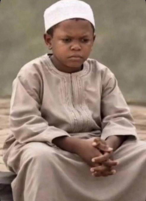 A young boy wearing a white cap and traditional light-colored robe sits with his hands clasped, looking serious.