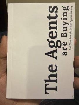 A hand holding a white book titled 'The Agents are Buying' with the subtitle 'Find Value For the Random Agents Economy' printed in black text.