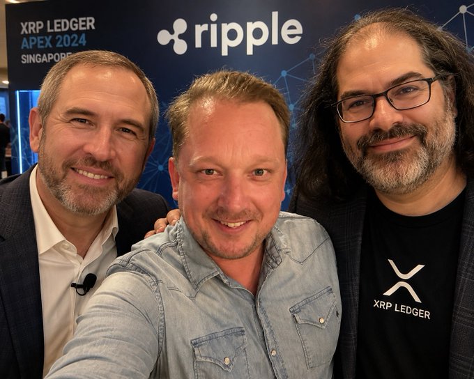 The image shows three men posing for a photo, with one of them wearing a black shirt that says "XRP Ledger" on it. The other two men are standing next to each other, smiling at the camera. They appear to be attending an event or gathering related to cryptocurrency and finance.