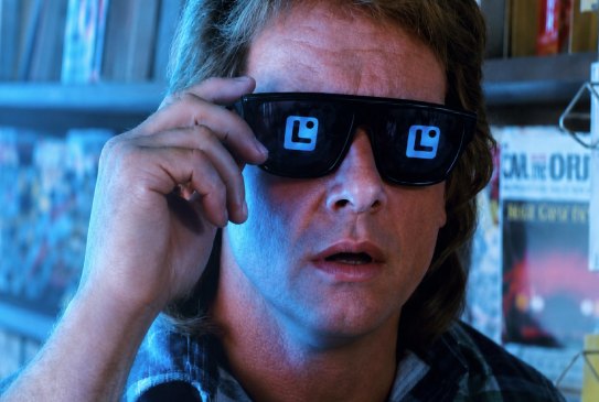 A man with wavy blonde hair puts on sunglasses that display digital emoticons in place of his pupils, with magazine covers visible in the background.