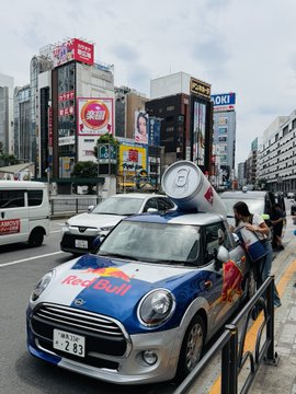 The image features a car with a can of Red Bull on top, driving down a city street surrounded by tall buildings. There are several people walking around the area, some carrying handbags or backpacks. The scene captures the bustling atmosphere of an urban environment where various elements coexist, including vehicles and pedestrians going about their daily routines.