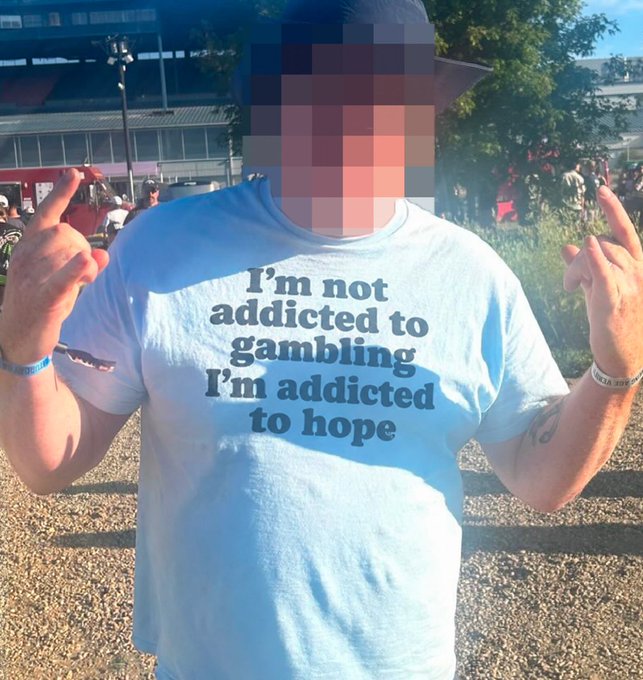 A person with a blurred face wearing a black cap and a white t-shirt that reads 'I'm not addicted to gambling I'm addicted to hope', holding up their middle fingers outdoors.
