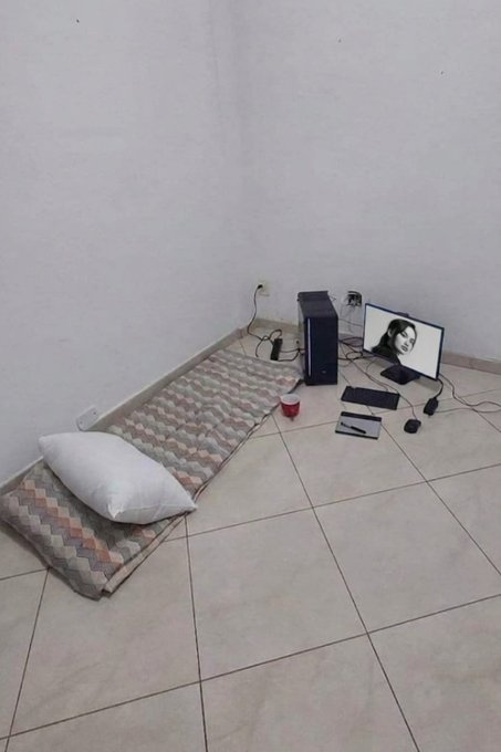 A sparse tiled room holds a thin mat, pillow, desktop computer setup, and a few scattered accessories, humorously illustrating a modest “lock-in” lifestyle funded by internet money. No text, numbers, tickers, or names are visible.