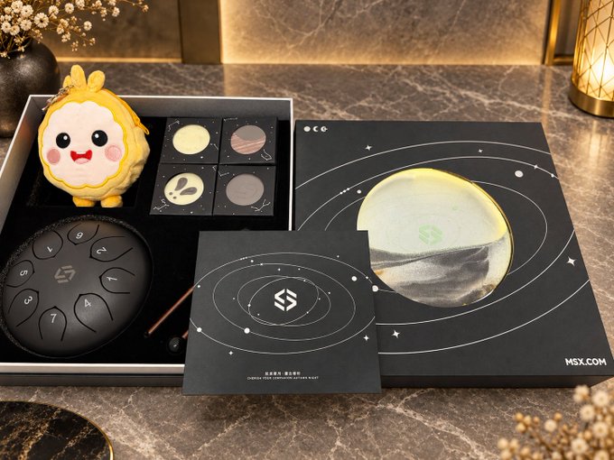 A black gift box set on a marble surface featuring space-themed chocolates, a moon-shaped item, a circular device with numbers, and a small yellow character figurine, with MSX.COM branding visible.