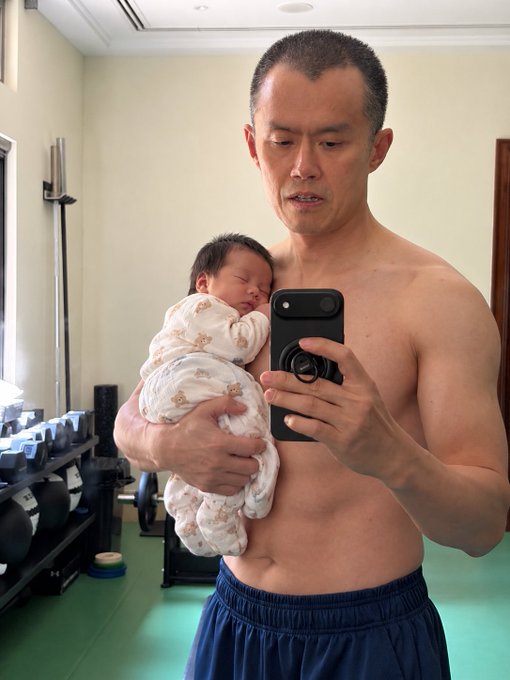 A shirtless man takes a mirror selfie in a gym while holding a sleeping baby wrapped in a teddy bear-patterned blanket.