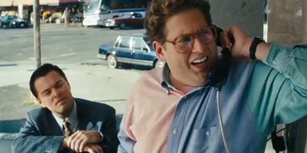 Film still showing Jonah Hill on a phone call wearing a pink and blue shirt and glasses, with Leonardo DiCaprio seated behind him in a suit.
