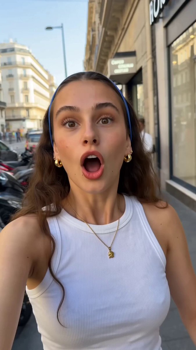 The image features a woman with long brown hair wearing a white tank top and a blue headband. She appears surprised as she takes a selfie using her phone's camera. The photo captures her face, which is the main focus of the image. There are no other people or objects in the scene, making it a simple yet engaging snapshot.