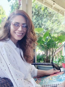 A smiling woman with glasses sits at an outdoor patio table surrounded by lush greenery, celebrating the launch of her NFT art collection titled "When the moon took root" with a Saturday auction starting at 0.03 ETH on @TransientLabs.