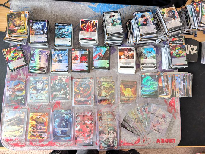 A collection of Azuki TCG cards fanned out and stacked on an Azuki playmat, showcasing various hits and holos pulled from presale boxes, with the "AZUKI" logo visible on the mat's bottom right corner.