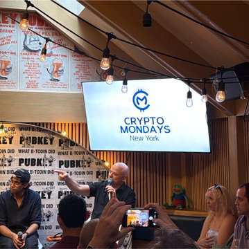 A panel discussion takes place at a "Crypto Mondays New York" event, with a man speaking into a microphone on a stage backed by a screen displaying the event's logo, while attendees capture the moment on their phones amid a festive atmosphere with string lights overhead.