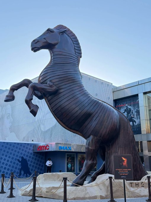 A massive rearing horse statue stands outside an AMC IMAX theater, where a plaque identifies it as the "Dark Horse" from "Mad Max: Fury Road" and a "Horse of the Apocalypse." The towering, stylized sculpture—with its armored, ribbed body—looks far more like a medieval knight's steed than an actual horse, fitting the post's playful jab at Christopher Nolan.