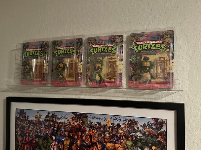 Four sealed Teenage Mutant Ninja Turtles action figure blister packs are displayed on a clear wall shelf, positioned above a framed Marvel comic poster signed by Stan Lee and other artists.