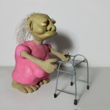 A whimsical claymation-style figurine of a hunched, elderly creature with sparse white hair and a pink dress, using a metal walker on a plain surface.