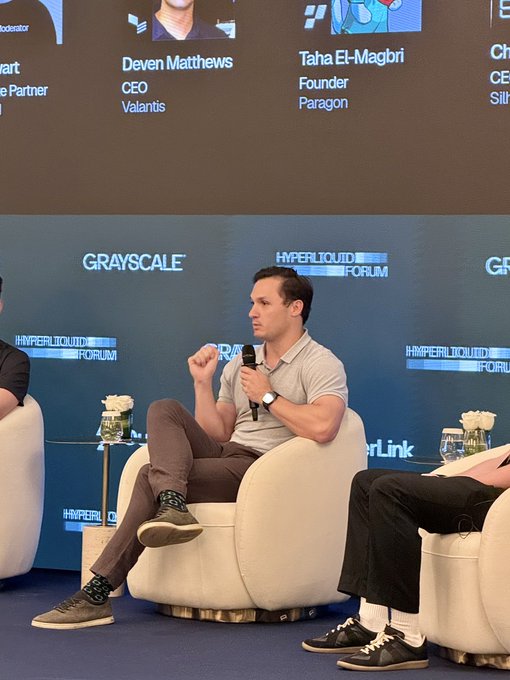 Deven Matthews, CEO of Valantis, speaking at the Hyperliquid Forum while seated on a white chair, with other panelists partially visible and a screen showing 'Taha El-Magbiri, Founder, Paragon' in the background.