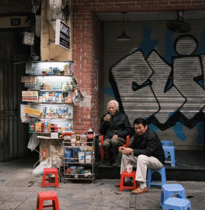 A street photography scene of two men sitting on small plastic stools outside a shop with a metal shutter covered in graffiti; one elderly man drinks from a glass while the other sits with hands clasped, surrounded by various bottled goods and a teapot.