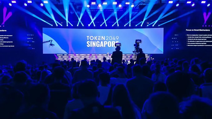 A large conference hall with blue stage lighting and a screen displaying 'TOKEN2049 SINGAPORE' to an audience of attendees.