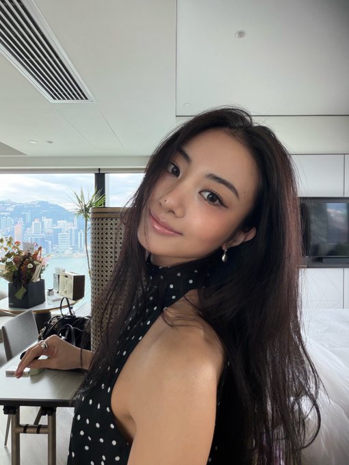 A selfie of a young woman with long dark hair wearing a black and white polka dot halter dress in a modern room with a city skyline view through the window.