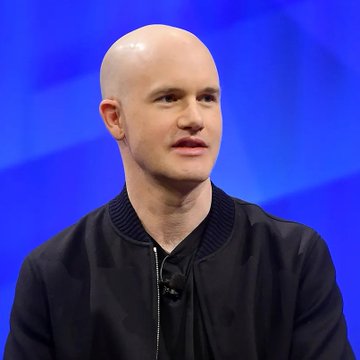 A bald man in a black bomber jacket is speaking at what appears to be a conference with a blue backdrop. The image is associated with Coinbase CEO Brian Armstrong's statement that the SEC and CFTC can establish clear cryptocurrency regulations under existing authority.