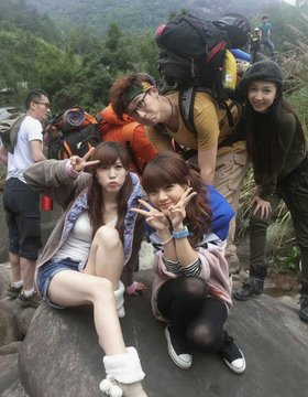 A group photo of Asian hikers/tourists posing on a rocky mountain summit, with two young women in the foreground making peace signs while others with backpacks smile behind them.