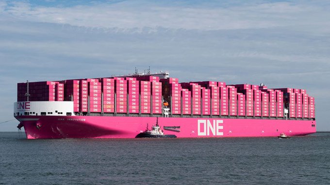 A large pink ONE container ship loaded with magenta containers sails on the ocean, accompanied by a small tugboat.