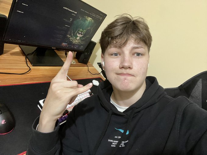 A young person wearing a black hoodie sits in front of a computer monitor displaying a Bear Grills game, making a peace sign with their hand.