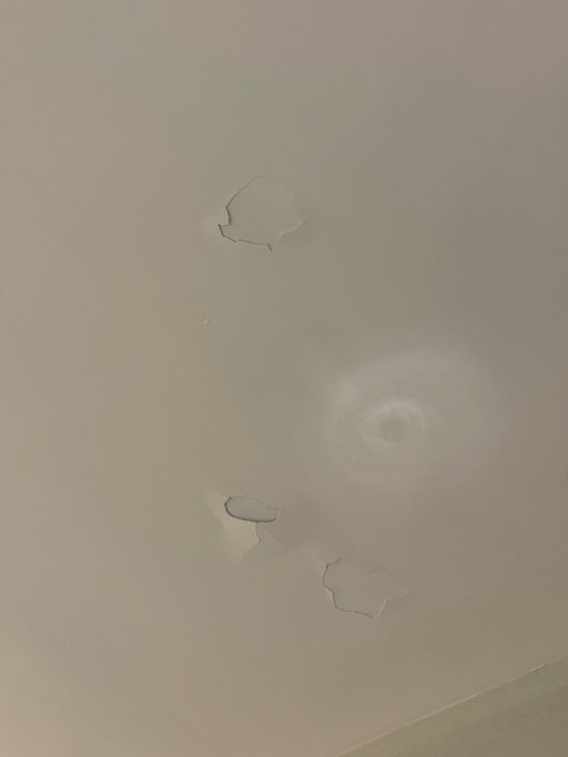 A close-up photo of a beige ceiling showing water damage with peeling paint and a damp circular stain.