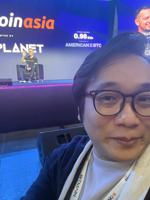 A man with glasses takes a selfie in the front row center of what appears to be the Binance Asia conference, with CZ visible on stage in the background and the event's "oinasia" branding, "0.96 ZNS" sponsorship numbers, and "AMERICAN BTC" text displayed on the large screen behind him. The post's caption "Hi, CZ" and joke about being noticed from his front-row center seat align perfectly with the image, showing both the attendee and the Binance founder speaking at the panel on stage.