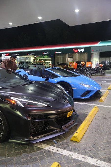 Two high-performance sports cars—a matte black and a bright blue Lamborghini—park at a gas station, likely during a road trip or cruise. A man in a red shirt and black cap is seen inside the dark car, while the post's message suggests a playful or flirtatious exchange between the two drivers.