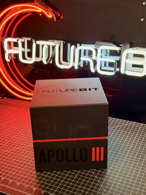 The image features a black box with red text that reads "Apollo III" and "We don't build miners. We build the best freedom tool on the Bitcoin network". The box has a unique design, resembling a watch face or a wristwatch. It is placed on a table next to a neon sign that says "futurebit". The neon sign is red with white text, which reads "futurebit" and "Apollo III", indicating that this box contains an item related to Bitcoin mining.