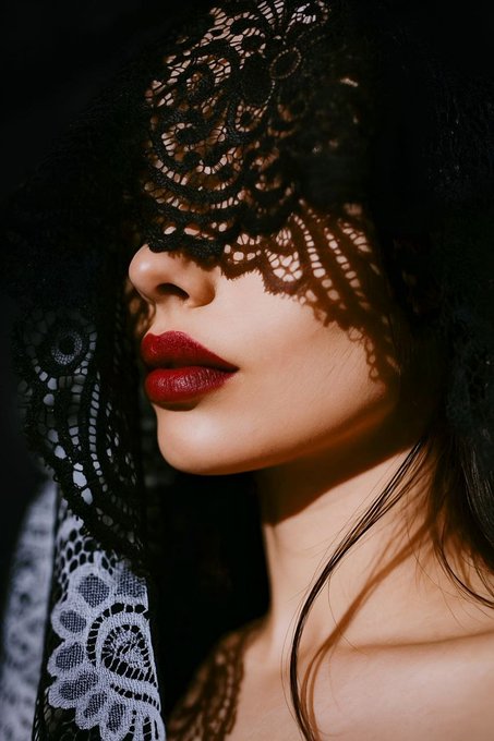 A woman with bold red lips poses under an intricate black lace veil, her face partially obscured while warm light highlights her cheekbone and lips against the dark shadows. The image embodies the mysterious, enchanting mood of the accompanying NFT post, where the delicate lacework and dramatic contrast evoke the theme of beauty emerging from darkness.