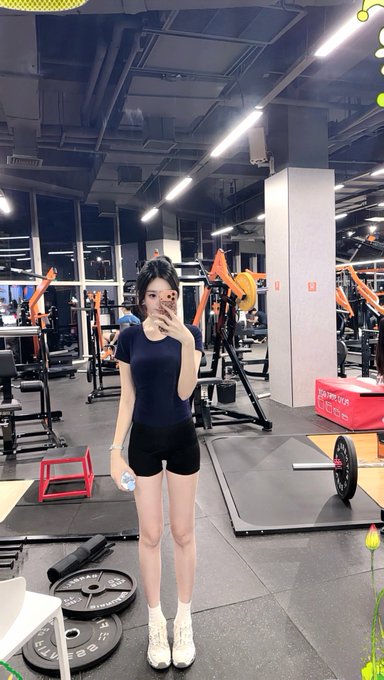 Mirror selfie of a young woman in a black t-shirt and shorts standing in a gym with various workout equipment visible in the background.