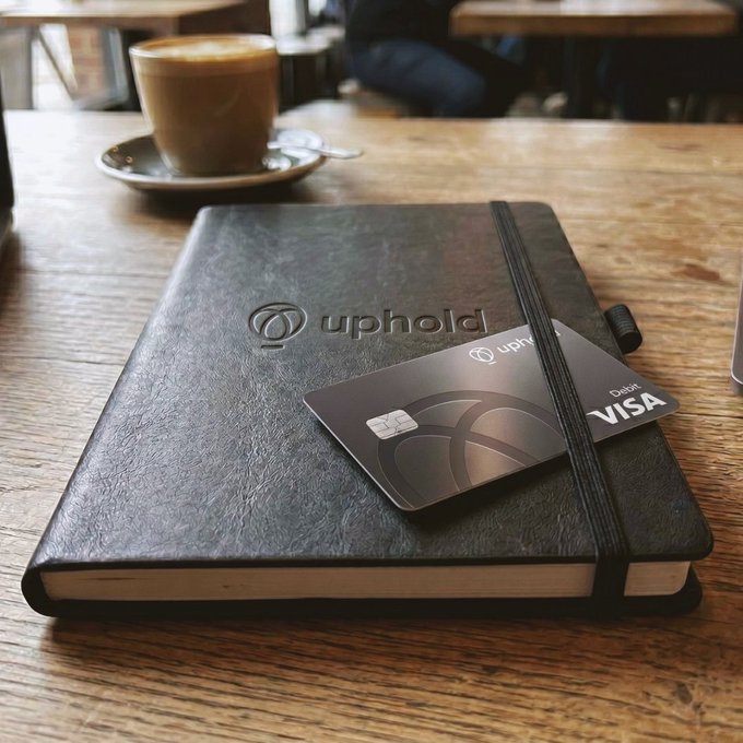 A black Uphold-branded notebook with an embossed logo sits on a wooden café table alongside an Uphold Debit Visa card and a cappuccino, capturing a relaxed weekend moment that ties perfectly to earning XRP rewards on the Uphold platform.