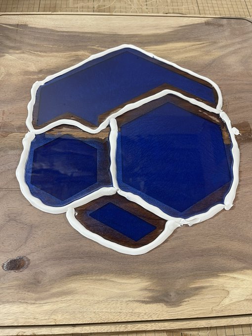 A custom-shaped resin art piece featuring deep blue geometric designs bordered by white resin and natural wood, resting on a wooden surface.