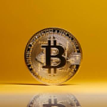 A golden Bitcoin (₿) cryptocurrency coin standing upright on a reflective yellow surface, with its reflection visible below.