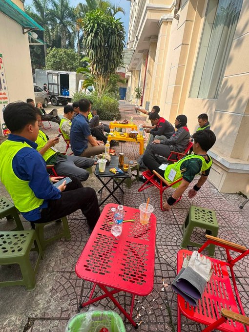 A group of men in work uniforms sit on small plastic chairs on a sidewalk in Vietnam, drinking and relaxing around a red plastic table.