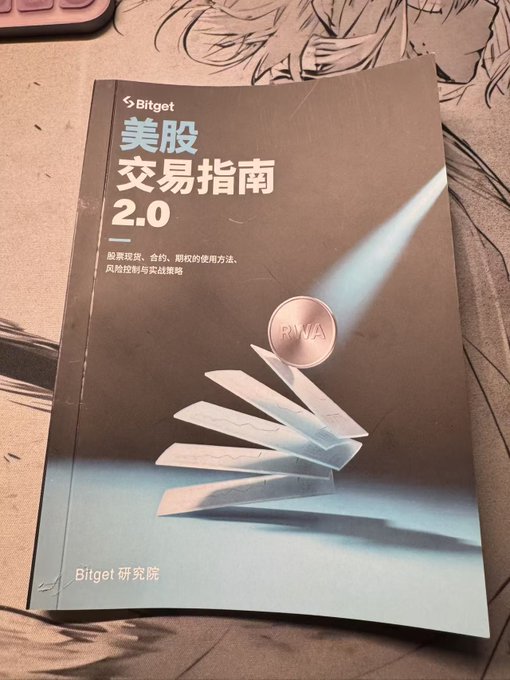A Bitget "美股交易指南 2.0" (US Stock Trading Guide 2.0) booklet published by the Bitget Research Institute (Bitget 研究院), featuring an RWA coin graphic, sits on an illustrated desk mat—promoting Bitget's trading education resources alongside the company's recent announcement of compensating users for approximately $40 million in losses from anomalous market conditions.