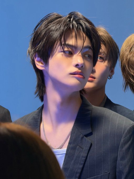 A close-up of a young man with dark, slightly messy hair wearing a gray pinstripe blazer over a white top and silver chain, with another person partially visible behind him against a blue background.